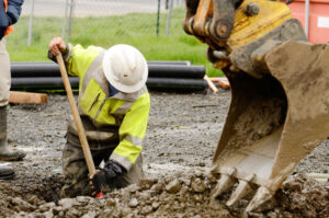 Worker using a small, tracked excavator to dig a hole to fix a water leak at a large commercial housing development in the muddy spring weather.